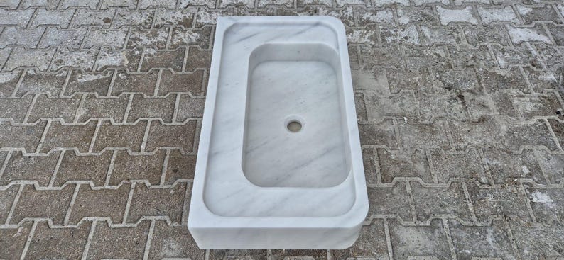 May include: A white marble sink with a rectangular basin and a smaller, offset rectangular area. The sink has a rounded edge and a drain hole in the center of the basin. The sink is made of white marble with gray veining.