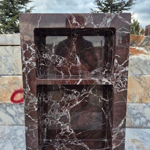 May include: A rectangular, dark red marble shower niche with two recessed shelves. The marble has a prominent white veining pattern. The niche is designed to be built into a shower wall for storage of toiletries.