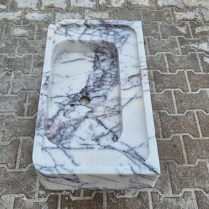 May include: A rectangular marble sink with a rounded interior basin. The sink is white with dark gray veining. A circular drain hole is visible at the bottom of the basin. The sink is set on a gray brick surface.