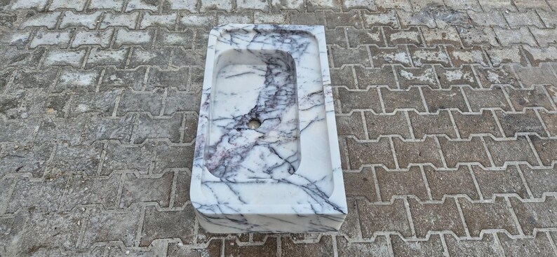May include: A rectangular marble sink with a rounded interior basin. The sink is white with dark gray veining. A circular drain hole is visible at the bottom of the basin. The sink is set on a gray brick surface.