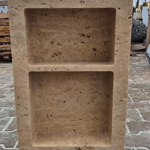 May include: A rectangular, beige stone structure with two recessed shelves. The stone has a textured surface with small holes and variations in color. The shelves are rectangular and evenly spaced, creating a symmetrical design. The structure is outdoors on a brick surface.