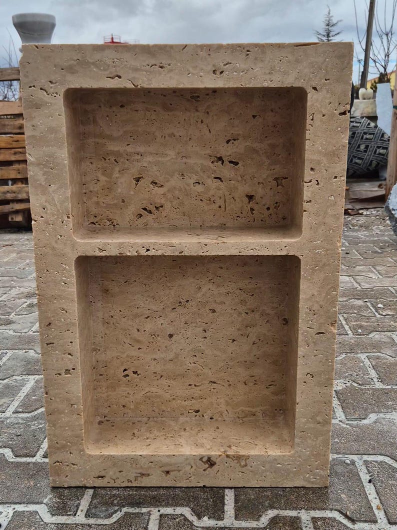May include: A rectangular, beige stone structure with two recessed shelves. The stone has a textured surface with small holes and variations in color. The shelves are rectangular and evenly spaced, creating a symmetrical design. The structure is outdoors on a brick surface.