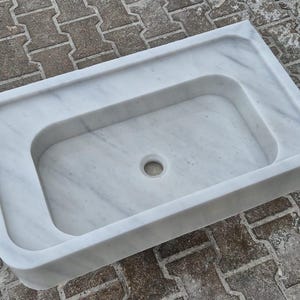 May include: A rectangular white marble sink with a central basin and a drain hole. The sink has a smooth, polished surface and a raised outer rim. The marble features subtle gray veining throughout. The sink is designed for bathroom use.