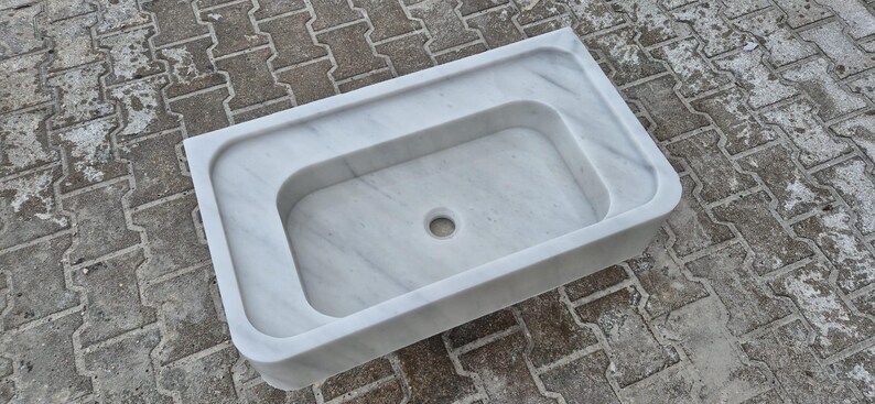 May include: A rectangular white marble sink with a central basin and a drain hole. The sink has a smooth, polished surface and a raised outer rim. The marble features subtle gray veining throughout. The sink is designed for bathroom use.