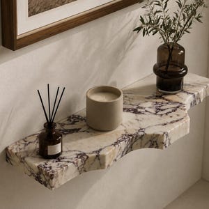 Puede incluir: Un estante de pared hecho de m&aacute;rmol blanco con vetas p&uacute;rpuras prominentes y un borde frontal curvo. Sobre el estante hay una peque&ntilde;a botella de vidrio marr&oacute;n con varillas difusoras negras, una vela beige cil&iacute;ndrica y un florero de vidrio oscuro.