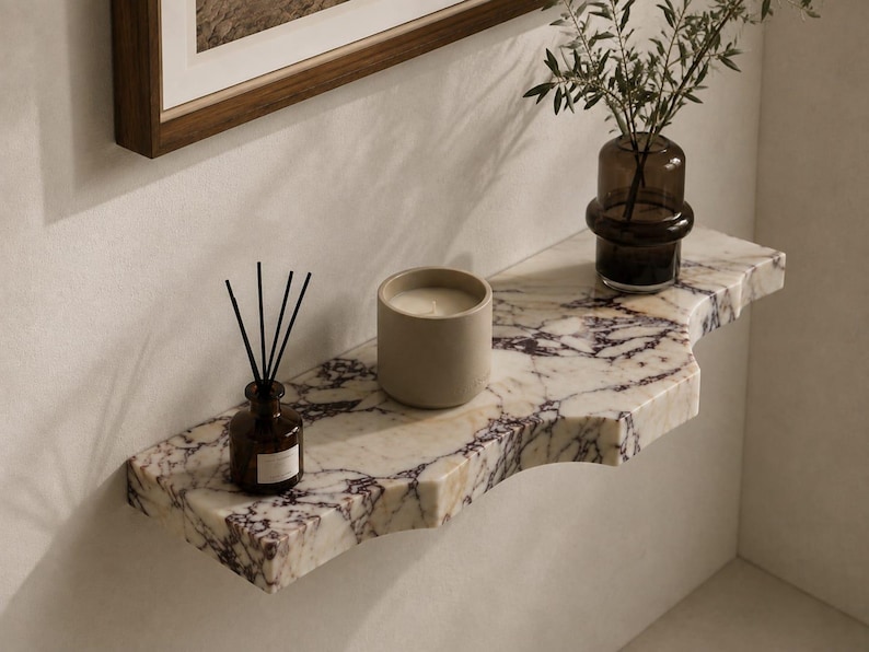 Puede incluir: Un estante de pared hecho de m&aacute;rmol blanco con vetas p&uacute;rpuras prominentes y un borde frontal curvo. Sobre el estante hay una peque&ntilde;a botella de vidrio marr&oacute;n con varillas difusoras negras, una vela beige cil&iacute;ndrica y un florero de vidrio oscuro.