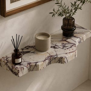 Puede incluir: Un estante de pared hecho de m&aacute;rmol blanco con vetas p&uacute;rpuras prominentes y un borde frontal curvo. Sobre el estante hay un peque&ntilde;o difusor de varillas de vidrio marr&oacute;n con varillas negras, una vela cil&iacute;ndrica beige y un florero de vidrio oscuro.