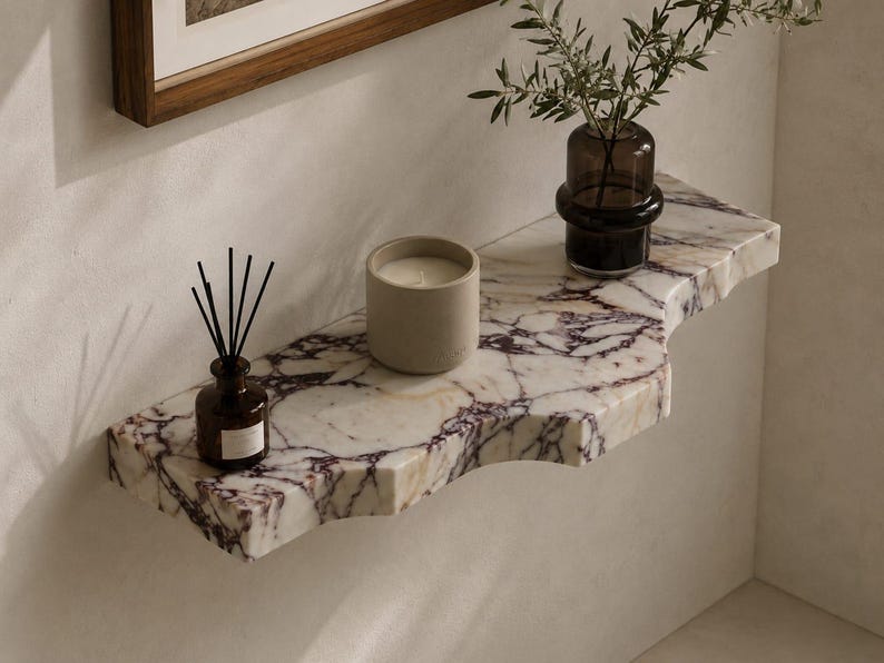 Puede incluir: Un estante de pared hecho de m&aacute;rmol blanco con vetas p&uacute;rpuras prominentes y un borde frontal curvo. Sobre el estante hay un peque&ntilde;o difusor de varillas de vidrio marr&oacute;n con varillas negras, una vela cil&iacute;ndrica beige y un florero de vidrio oscuro.