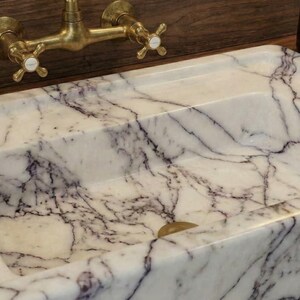 May include: A rectangular marble sink with a white and purple veined pattern. The sink has a brass faucet with cross handles. The sink is set on a wooden surface.