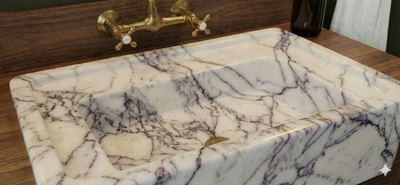 May include: A rectangular marble sink with a white and purple veined pattern. The sink has a brass faucet with cross handles. The sink is set on a wooden surface.