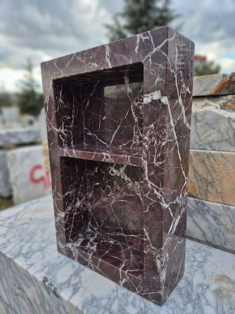 May include: A rectangular, freestanding marble shelf unit with two shelves. The marble is a deep burgundy color with white veining. The unit is designed to be recessed into a wall or used as a decorative storage piece.