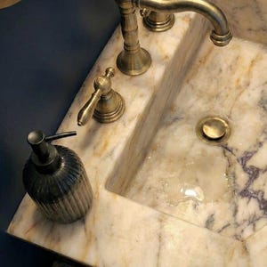 May include: A bathroom sink with a marble countertop and brass fittings. The sink has a built-in basin, a tap, and two handles. A dark, ribbed soap dispenser sits on the counter.
