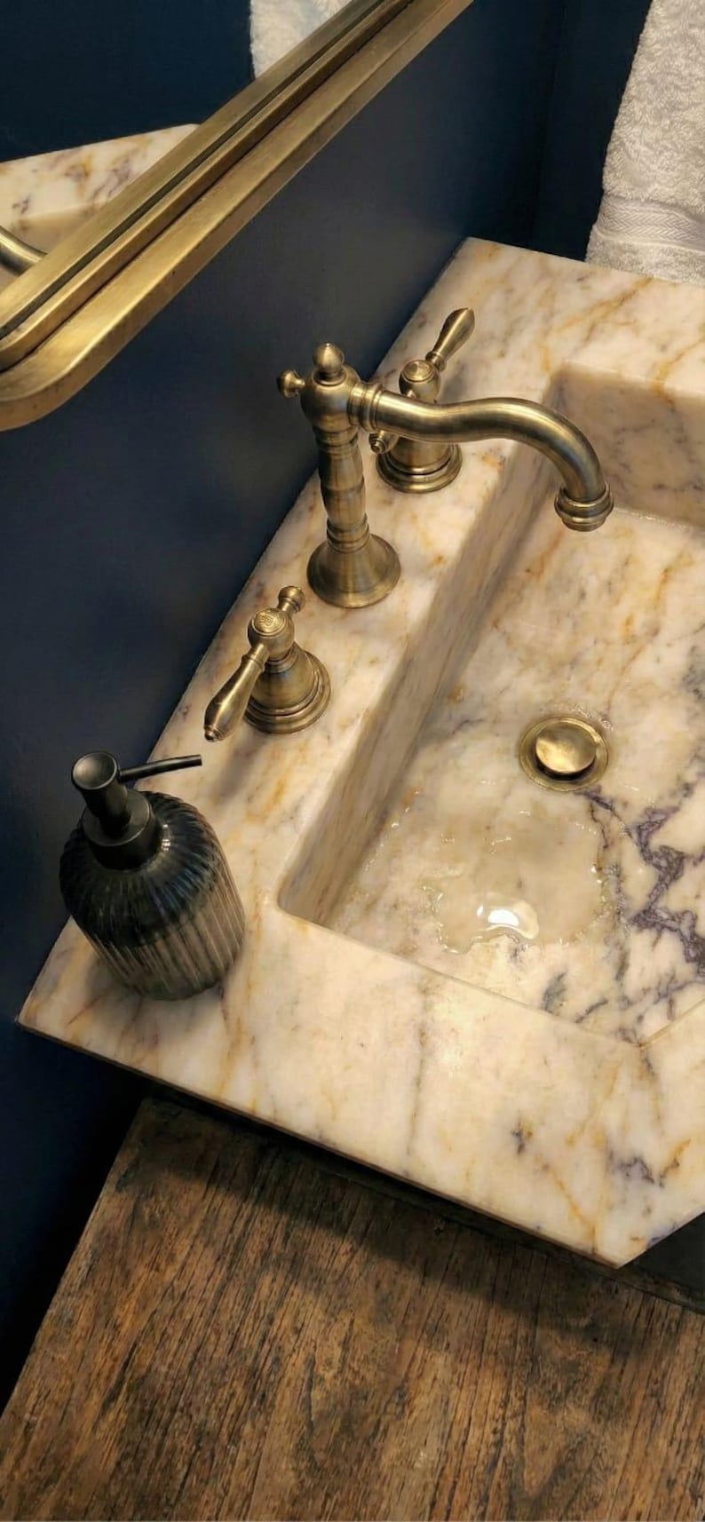 May include: A bathroom sink with a marble countertop and brass fittings. The sink has a built-in basin, a tap, and two handles. A dark, ribbed soap dispenser sits on the counter.