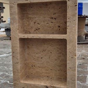 May include: A rectangular, beige stone shower niche with two shelves. The stone has a textured, porous surface. The niche is designed to be recessed into a wall for storage of bath products. The overall design is simple and functional.