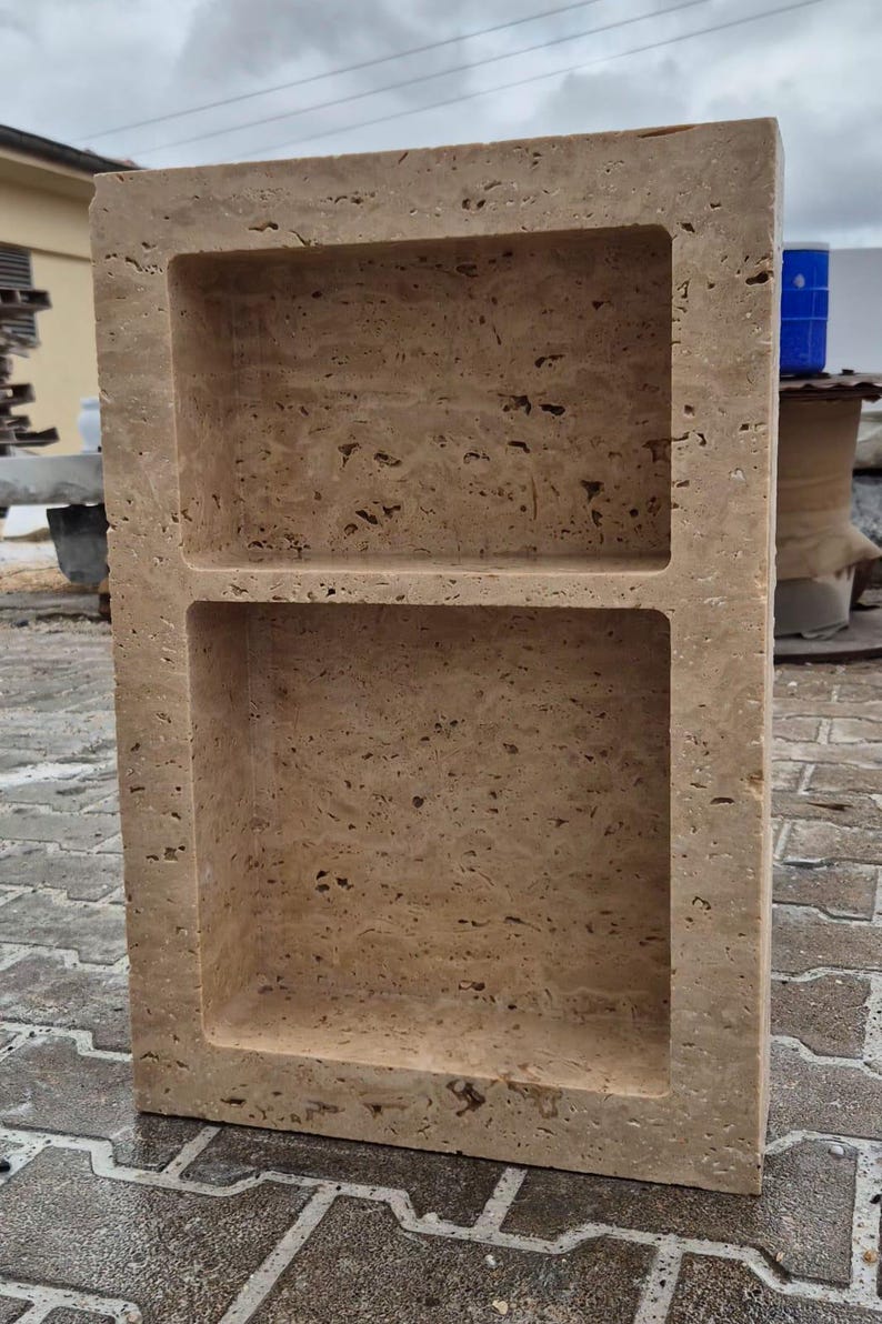 May include: A rectangular, beige stone shower niche with two shelves. The stone has a textured, porous surface. The niche is designed to be recessed into a wall for storage of bath products. The overall design is simple and functional.