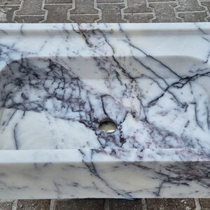 May include: Rectangular marble sink with a white base and dark gray veining. The sink has a rounded interior basin and a small drain hole. The sink is made of a solid block of marble.