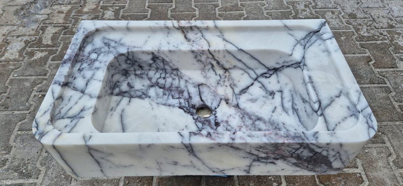 May include: Rectangular marble sink with a white base and dark gray veining. The sink has a rounded interior basin and a small drain hole. The sink is made of a solid block of marble.