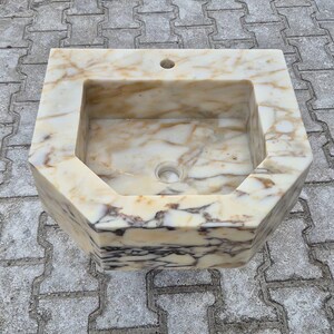 May include: An octagonal marble sink. The sink is a light beige colour with brown and white veining. It has a rectangular basin with a drain and a hole for a tap.