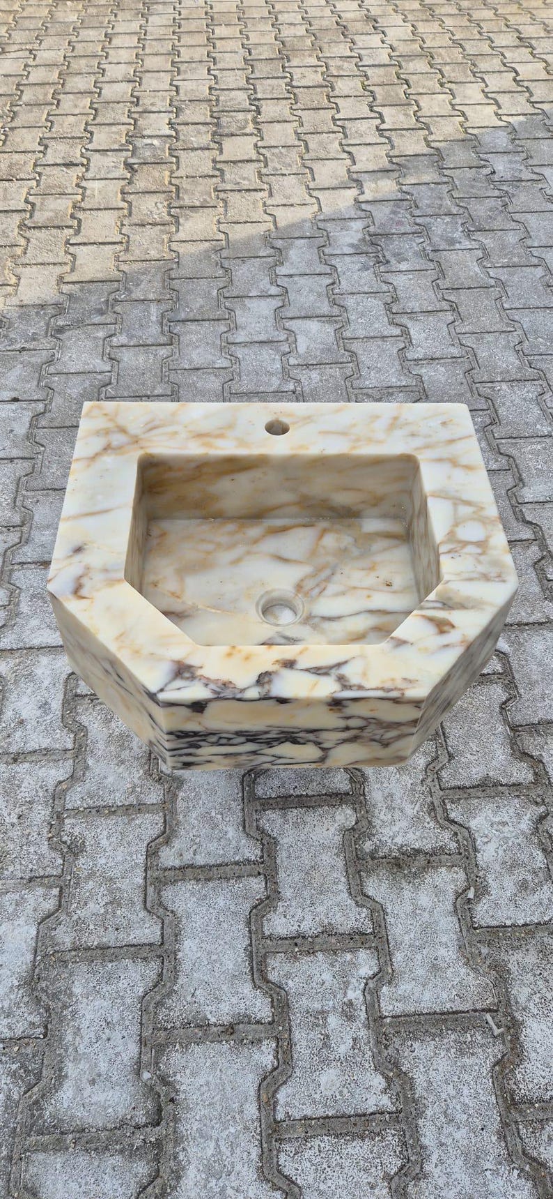 May include: An octagonal marble sink. The sink is a light beige colour with brown and white veining. It has a rectangular basin with a drain and a hole for a tap.