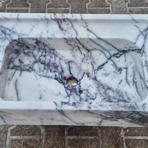 May include: Rectangular marble sink with a white base and dark gray veining. The sink has a rounded interior basin and a raised, flat area along the back edge. A drain hole is visible in the center of the basin.