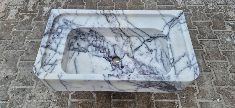 May include: Rectangular marble sink with a white base and dark gray veining. The sink has a rounded interior basin and a raised, flat area along the back edge. A drain hole is visible in the center of the basin.