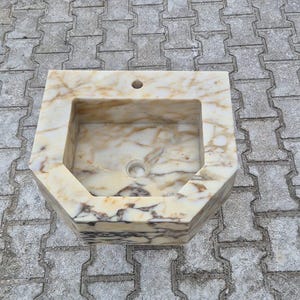 May include: A rectangular marble sink with a light beige and brown veined pattern. The sink has a square basin and an octagonal exterior shape. It features a drain hole and a faucet hole.