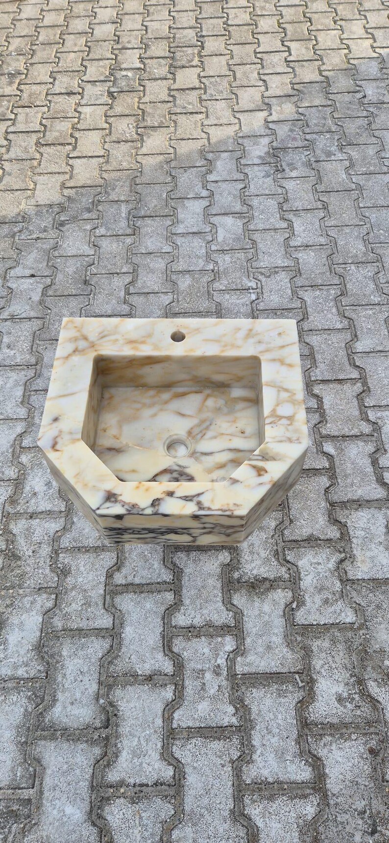 May include: A rectangular marble sink with a light beige and brown veined pattern. The sink has a square basin and an octagonal exterior shape. It features a drain hole and a faucet hole.