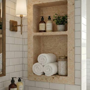 May include: A built-in bathroom shelf with two levels, holding rolled white towels, glass jars, and bottles of soap and lotion. A small potted plant adds a touch of greenery. The shelf is made of a light-colored stone and is set into a white tiled wall.