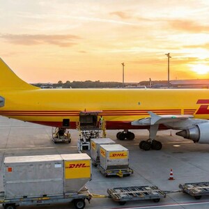 May include: A yellow DHL cargo plane on a tarmac at sunset. The plane has red stripes and the DHL logo. Cargo containers are being loaded onto the plane. The sky is a mix of orange and yellow.