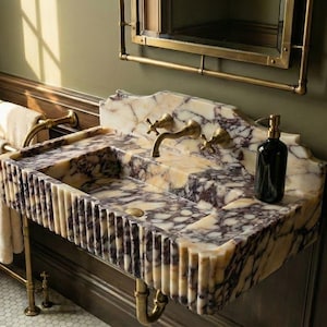 Pu&ograve; includere: Un lussuoso lavabo da bagno con piano in marmo con venature viola e crema. Il lavabo ha un bordo scanalato e rubinetti in ottone. Una bottiglia nera &egrave; appoggiata sul bancone.