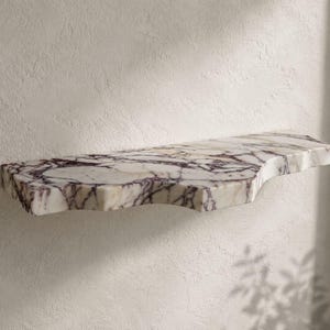 Puede incluir: Un estante de pared hecho de m&aacute;rmol blanco con vetas de color p&uacute;rpura oscuro irregulares y prominentes. El estante presenta una superficie lisa y pulida con una silueta org&aacute;nica y asim&eacute;trica de bordes curvos, montado horizontalmente en una pared texturizada.