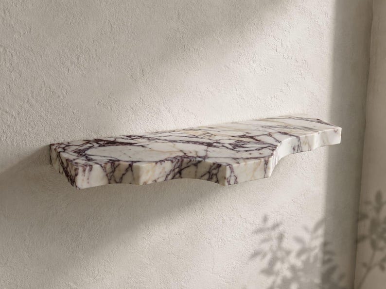 Puede incluir: Un estante de pared hecho de m&aacute;rmol blanco con vetas de color p&uacute;rpura oscuro irregulares y prominentes. El estante presenta una superficie lisa y pulida con una silueta org&aacute;nica y asim&eacute;trica de bordes curvos, montado horizontalmente en una pared texturizada.