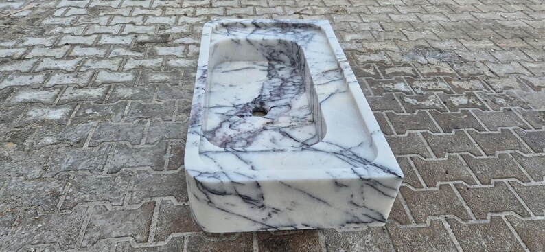 May include: A rectangular marble sink with a white base and dark gray veining. The sink has a rectangular basin with a drain hole. The sink is made of stone and is suitable for a bathroom.