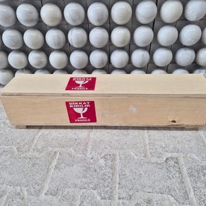 Puede incluir: Una caja rectangular de madera de color claro con dos pegatinas cuadradas rojas en la parte frontal. Cada pegatina muestra un icono de copa de vino blanca con el texto DIKKAT KIRILIR y FRAGILE en blanco, indicando que el contenido es fr&aacute;gil.