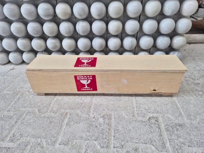 Puede incluir: Una caja rectangular de madera de color claro con dos pegatinas cuadradas rojas en la parte frontal. Cada pegatina muestra un icono de copa de vino blanca con el texto DIKKAT KIRILIR y FRAGILE en blanco, indicando que el contenido es fr&aacute;gil.