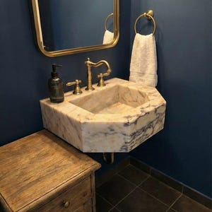 May include: A bathroom with a marble sink and brass fittings, a gold-framed mirror, and a wooden cabinet. The sink is mounted on a dark blue wall, with a white towel on a brass ring. The floor is tiled.