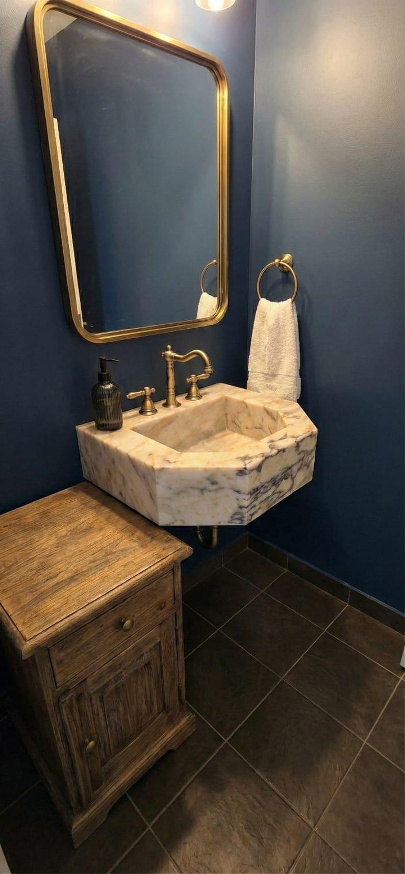 May include: A bathroom with a marble sink and brass fittings, a gold-framed mirror, and a wooden cabinet. The sink is mounted on a dark blue wall, with a white towel on a brass ring. The floor is tiled.
