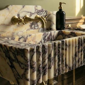 Pu&ograve; includere: Un lavabo da bagno in marmo con un rubinetto dorato e un dosatore di sapone scuro. Il lavabo presenta un design unico con scanalature verticali e una superficie a motivi color crema e viola. Il lavabo &egrave; sostenuto da gambe dorate.