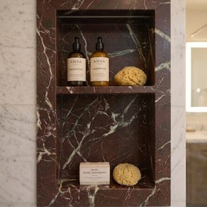 May include: A built-in marble shower niche with two shelves. The top shelf holds two bottles of "ANYA Shampoo" and a natural sponge. The bottom shelf has a bar of "ANYA Rose Geranium" soap and another sponge. The marble is a deep red with white veining.