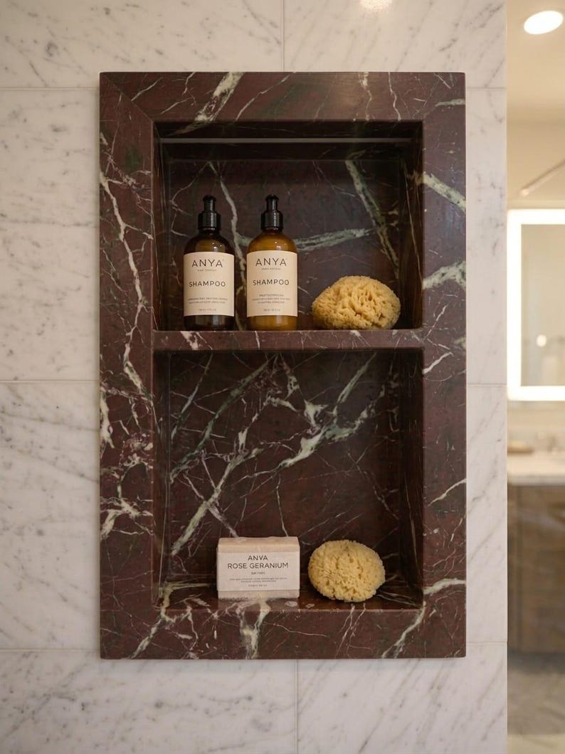 May include: A built-in marble shower niche with two shelves. The top shelf holds two bottles of "ANYA Shampoo" and a natural sponge. The bottom shelf has a bar of "ANYA Rose Geranium" soap and another sponge. The marble is a deep red with white veining.