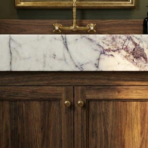 May include: A wooden bathroom vanity with a marble sink. The sink has purple veining. The vanity has two doors with brass knobs. A brass faucet and a black soap dispenser are visible. A white towel hangs on a brass ring.