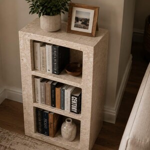 Pu&ograve; includere: Una libreria rettangolare in pietra travertino beige con tre ripiani aperti. I ripiani contengono vari libri con dorsi bianchi, neri e marroni, una piccola ciotola di legno e un vaso in ceramica. Una pianta e una foto incorniciata sono posizionate sopra.