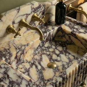 Pu&ograve; includere: Un lussuoso lavabo in marmo dal design unico. Il lavabo presenta un motivo venato color crema e viola, elementi dorati e un bordo scanalato. Sono visibili una bottiglia nera e un portasciugamani con asciugamani.