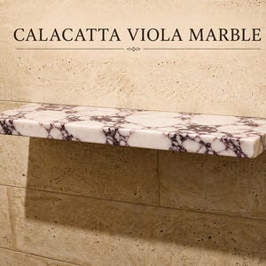 Può includere: Una mensola rettangolare da parete in marmo Calacatta Viola, caratterizzata da una base bianca con venature viola scuro. Il testo CALACATTA VIOLA MARBLE è stampato sopra la mensola, fissata su una parete con piastrelle beige.