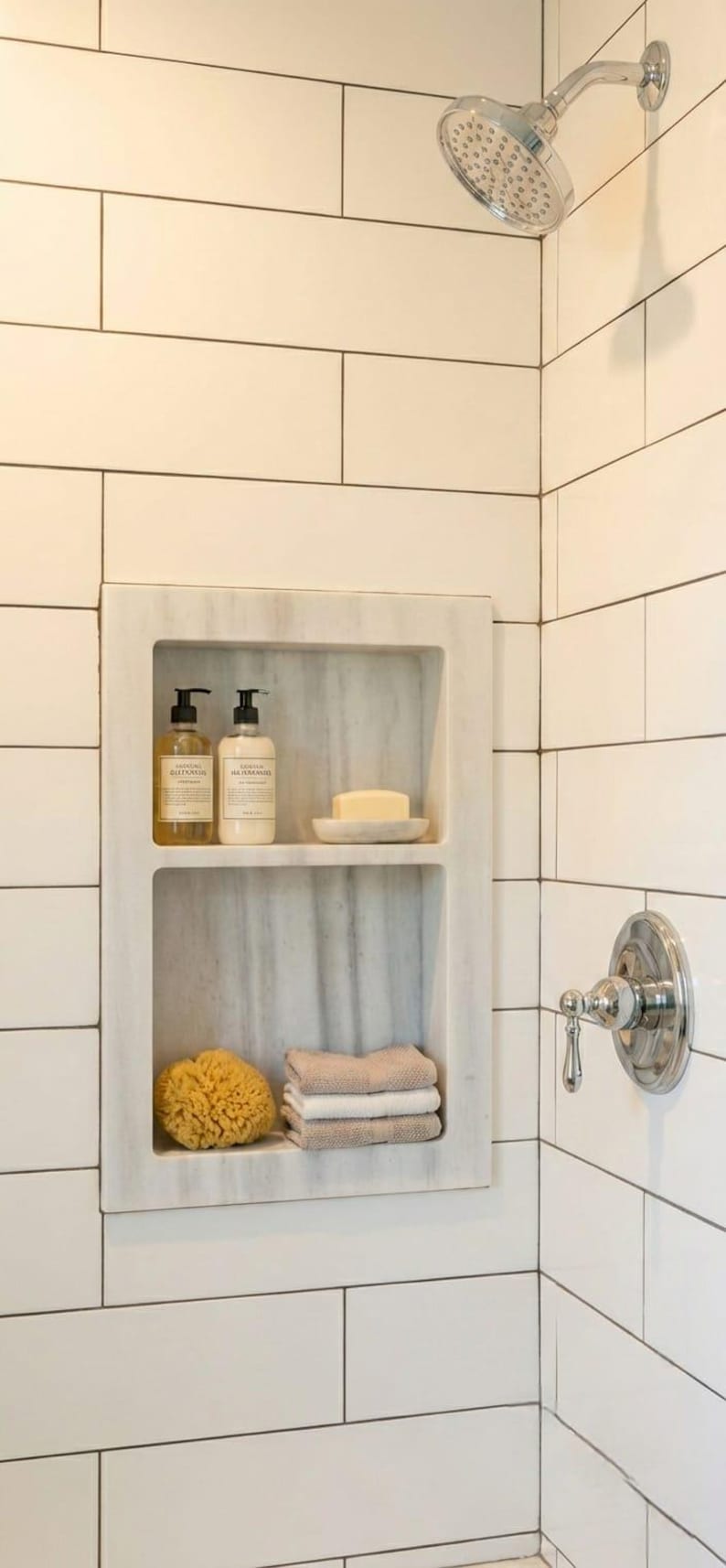 May include: A built-in marble shower niche with two pump bottles, a bar of soap, a natural sponge, and folded towels. The shower is tiled with white rectangular tiles and features a chrome shower head and faucet.
