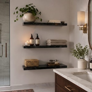 May include: Three black marble floating shelves mounted on a white wall in a bathroom. The shelves hold a potted plant, folded towels, two amber pump bottles, a candle, a woven box, and small glass jars. A vanity with a sink is nearby.