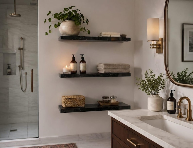 May include: Three black marble floating shelves mounted on a white wall in a bathroom. The shelves hold a potted plant, folded towels, two amber pump bottles, a candle, a woven box, and small glass jars. A vanity with a sink is nearby.