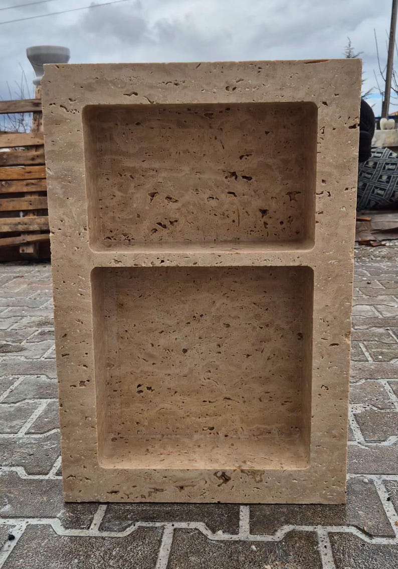 May include: A rectangular, beige stone shelf with two recessed compartments. The stone has a textured, porous surface. The shelf is freestanding and appears to be made of natural stone. The shelf is designed for storage or display.