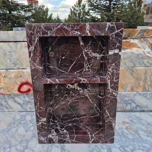 May include: A rectangular, burgundy marble shower niche with two recessed shelves. The marble has white veining throughout. The niche is displayed outdoors, with trees and other marble slabs in the background.
