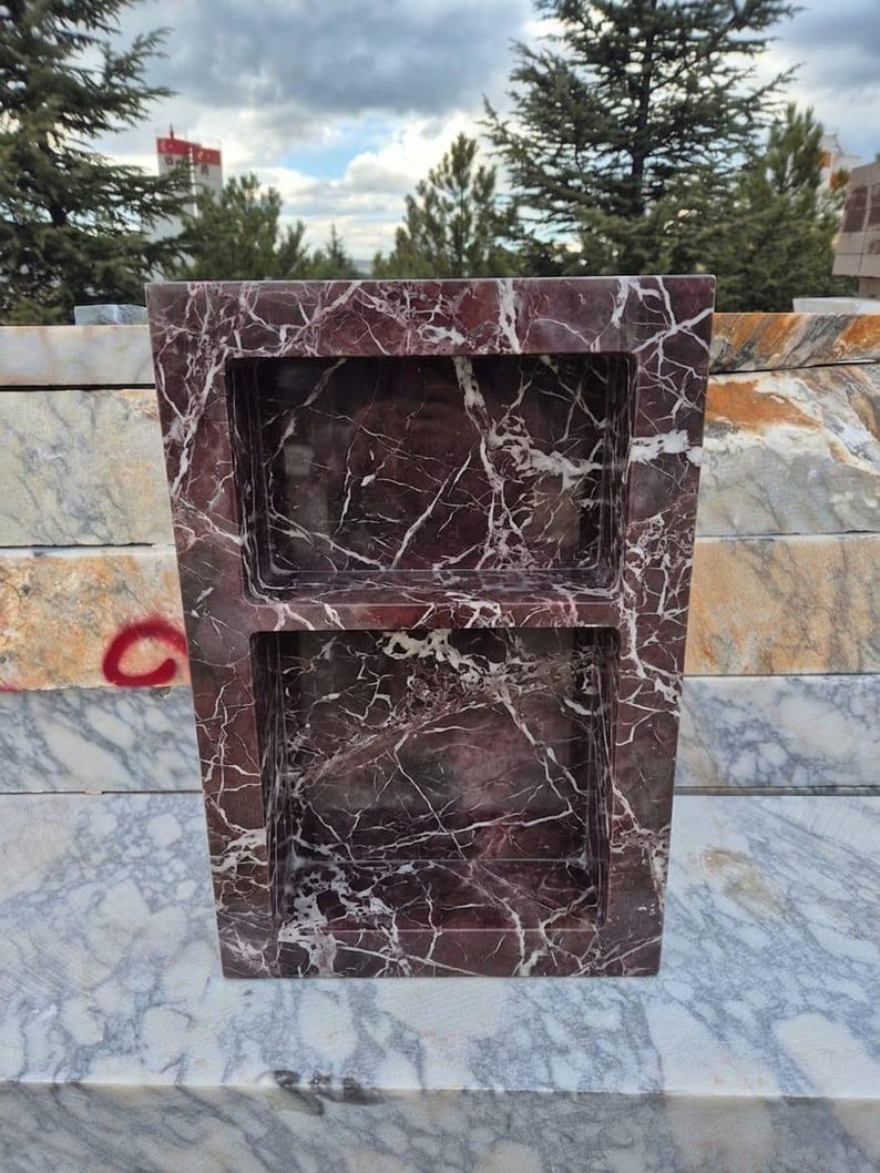 May include: A rectangular, burgundy marble shower niche with two recessed shelves. The marble has white veining throughout. The niche is displayed outdoors, with trees and other marble slabs in the background.